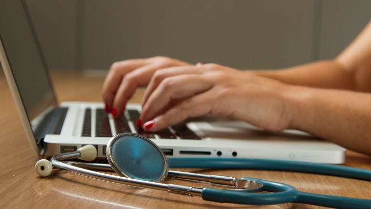 Image shows female hands typing on a laptop with a stethoscope next to the laptop